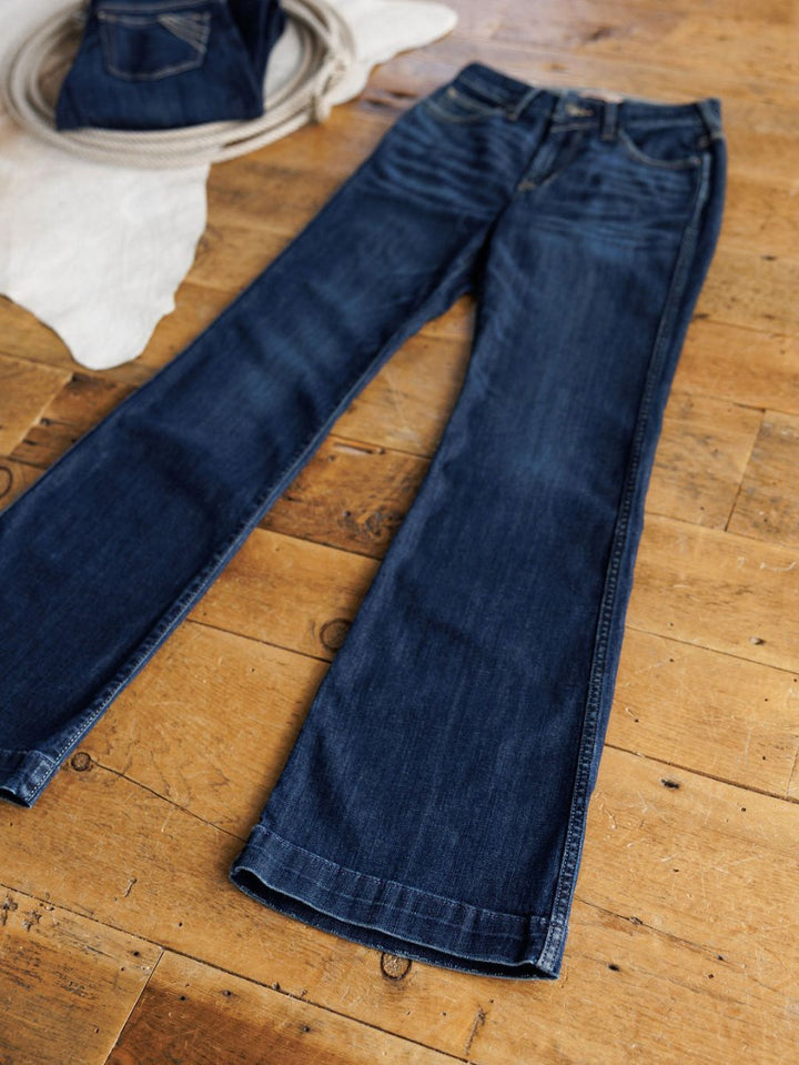 A pair of Ariat The Coy High Rise Slim Trouser Jeans in medium dark wash denim lies flat on a wooden floor, with another folded pair and a coiled rope visible in the background.