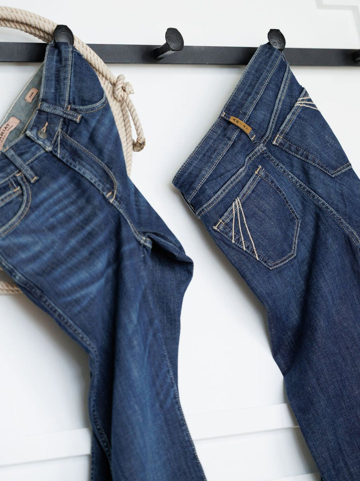 Two pairs of Ariat The Coy High Rise Slim Trouser Jeans hang on black wall hooks, with a beige rope draped over one hook, set against a white wall background.