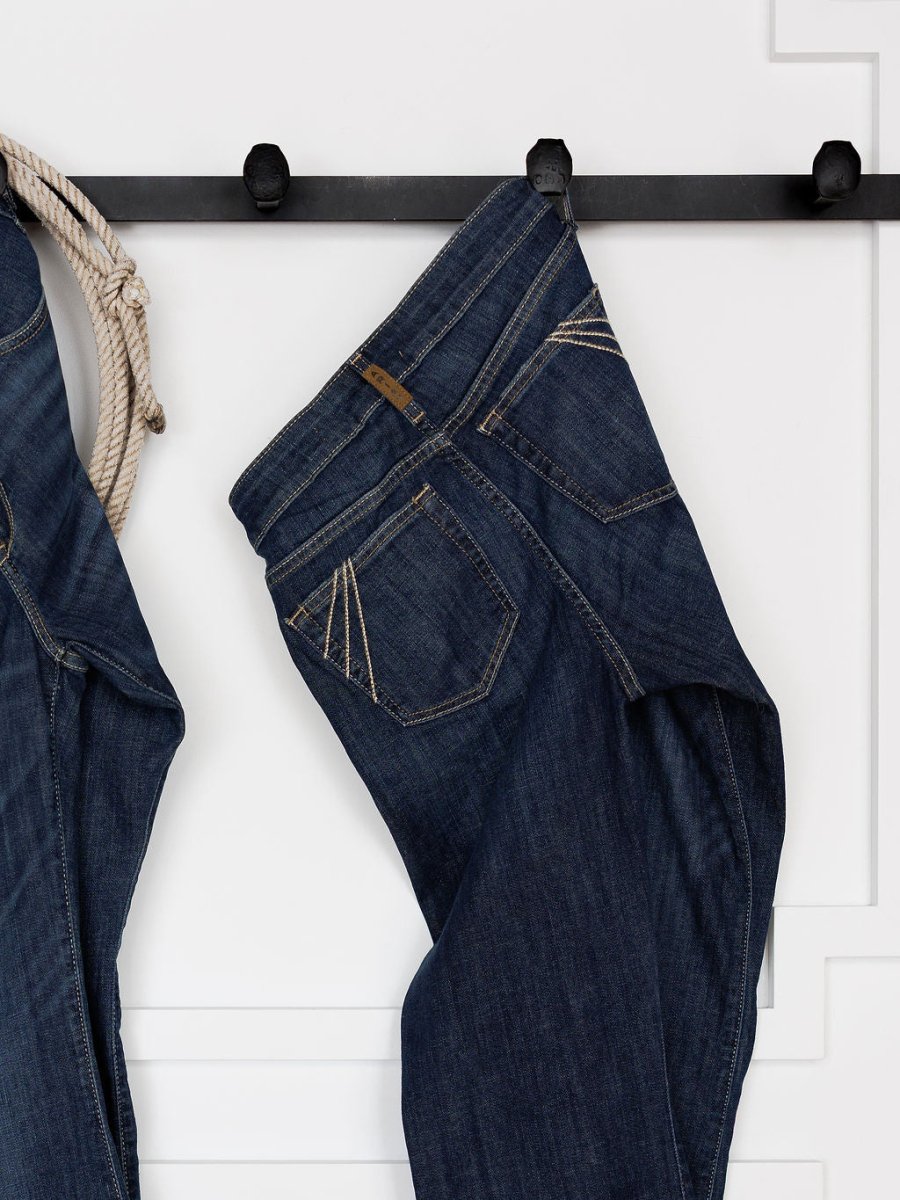 The Ariat Coy High Rise Slim Trouser Jean in a medium dark wash hangs from a black wall hook beside another pair of jeans and a rope, set against a white wall with decorative molding.