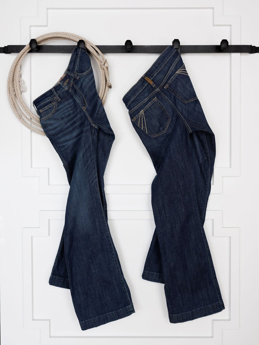 Two pairs of Ariat's The Coy High Rise Slim Trouser Jeans in medium dark wash hang side by side on a black wall rack against a white paneled background, with a coiled rope draped over the left end of the rack.