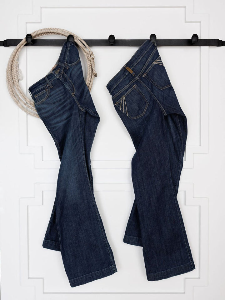 Two pairs of Ariat's The Coy High Rise Slim Trouser Jeans in medium dark wash hang side by side on a black wall rack against a white paneled background, with a coiled rope draped over the left end of the rack.