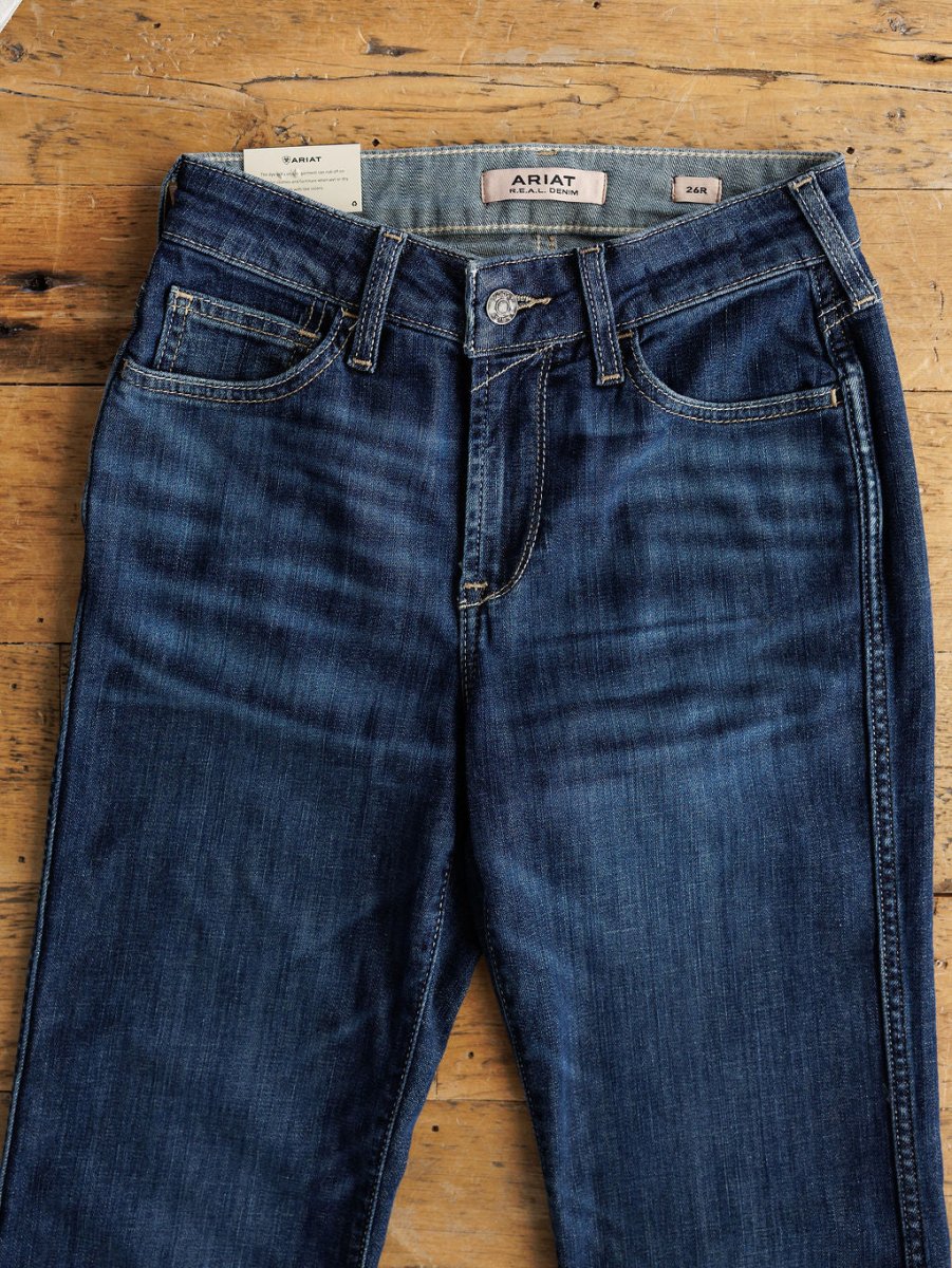 A pair of Ariat The Coy High Rise Slim Trouser Jeans in medium dark wash denim with button and zipper closure, front pockets, and visible stitching, laid flat on wood. Inner label reads ARIAT; size label shows 24M.