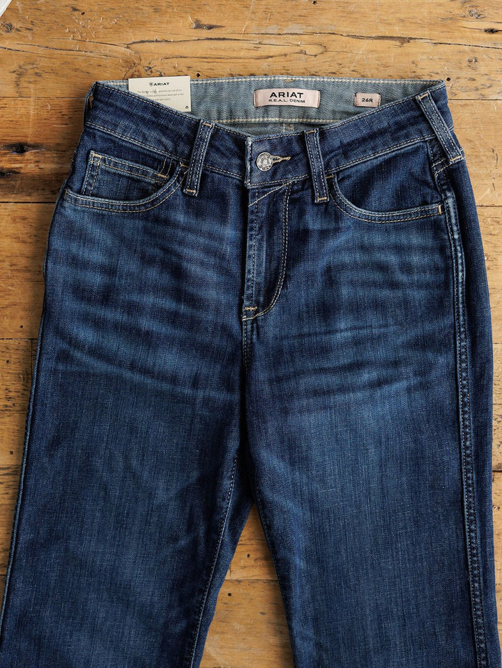 A pair of Ariat The Coy High Rise Slim Trouser Jeans in medium dark wash denim with button and zipper closure, front pockets, and visible stitching, laid flat on wood. Inner label reads ARIAT; size label shows 24M.