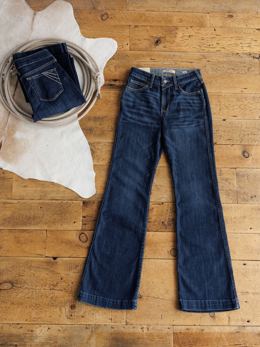 A pair of blue jeans lies flat on a wooden floor. Next to them, a folded pair of Ariat The Coy High Rise Slim Trouser Jeans in medium dark wash rests on white cowhide, accompanied by a coiled rope.