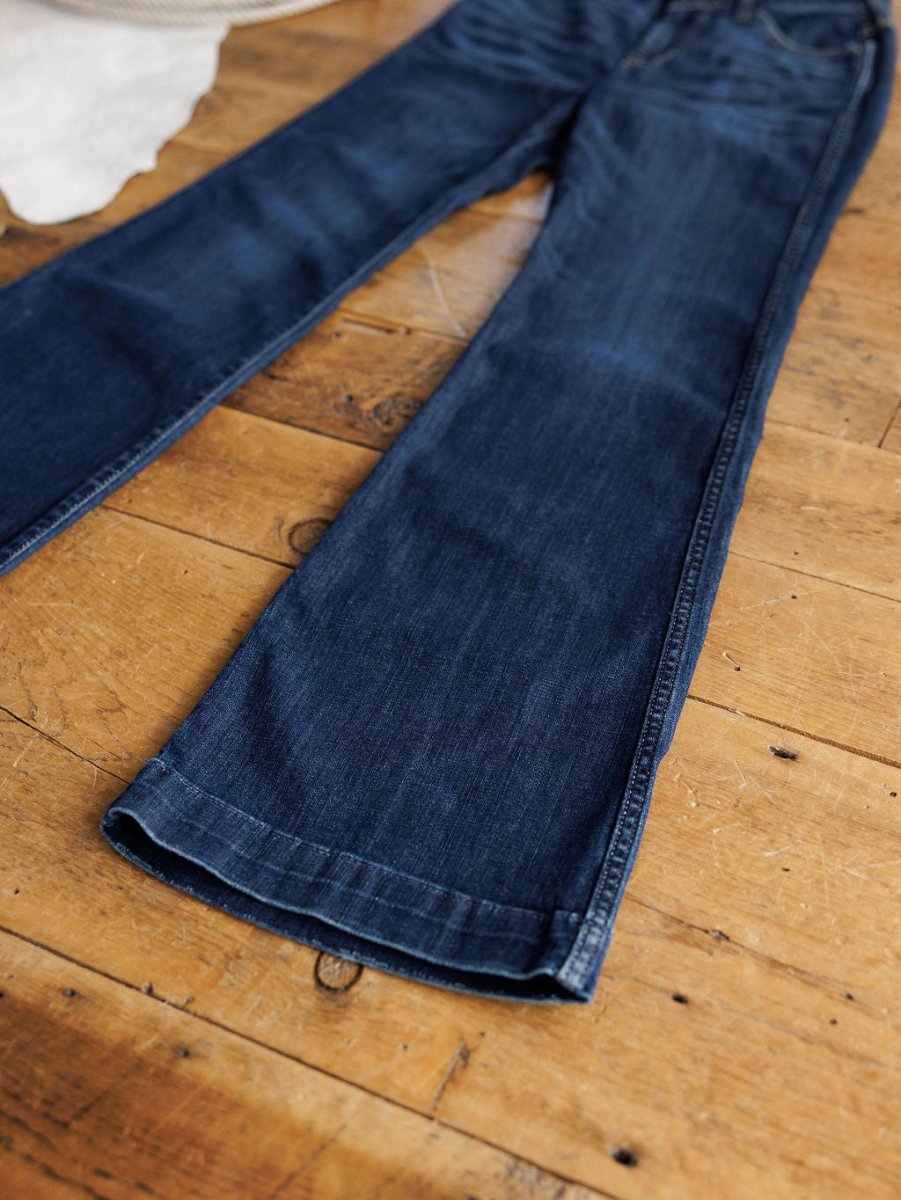 A pair of Ariat The Coy High Rise Slim Trouser Jeans in a medium dark wash with slim fit and flared legs is laid flat on a wooden floor.