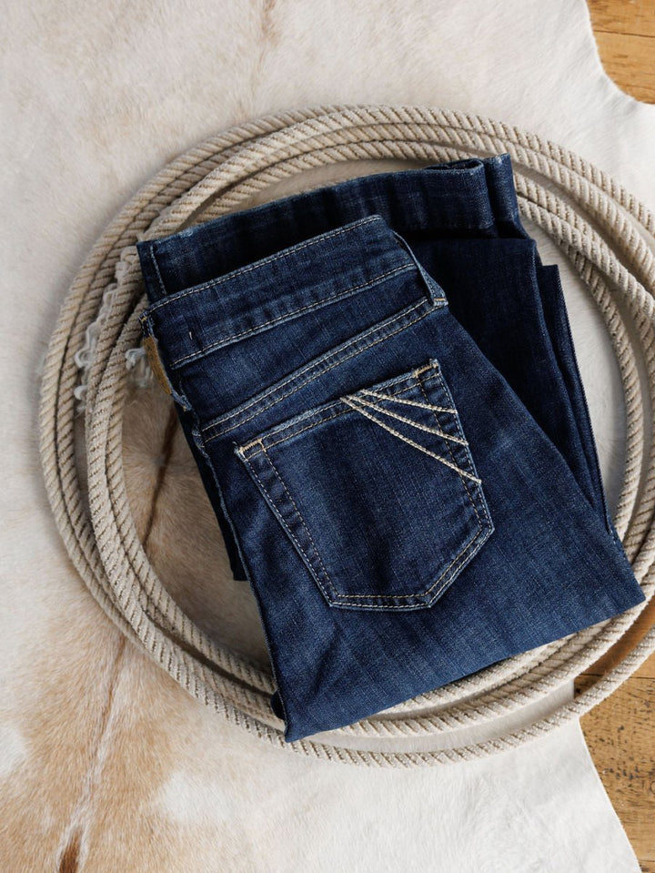 The Ariat Coy High Rise Slim Trouser Jean in medium dark wash denim rests on a coiled rope atop a light brown and white animal hide rug.