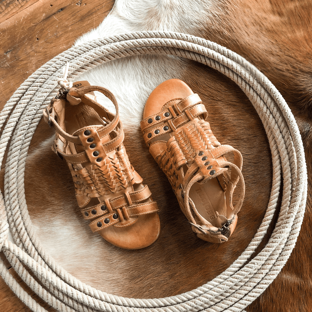 The Dacono tan woven leather sandals by Savannah Sevens western life{&}style, featuring studded details, rest on a coiled rope atop a cowhide rug and wooden floor. Sandals by BedStu. 