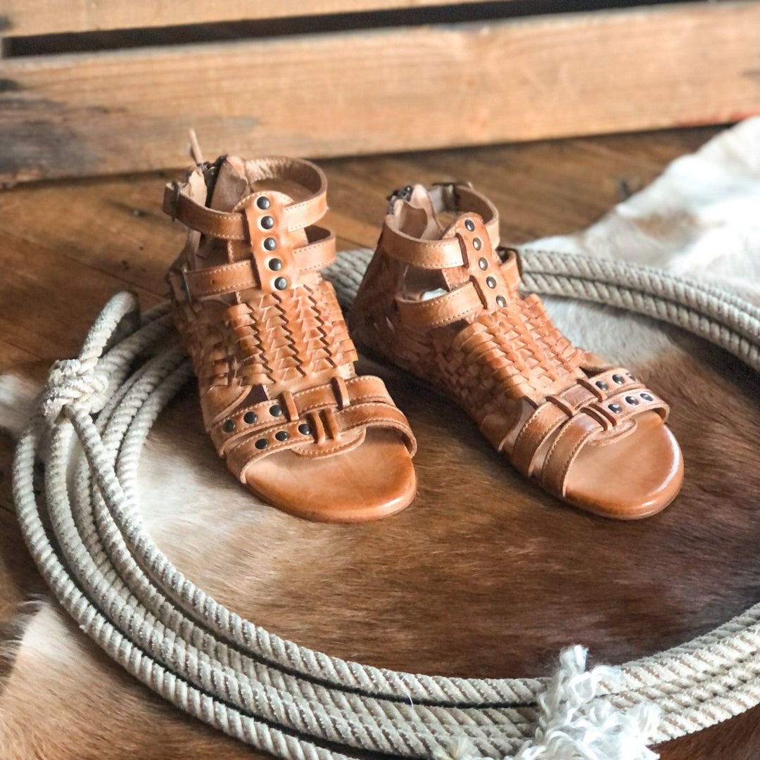 The Dacono tan woven leather flat sandals with metal studs by Savannah Sevens western life{&}style rest on a coiled rope, atop a cowhide rug and wooden floor. Sandals made by BedStu. 