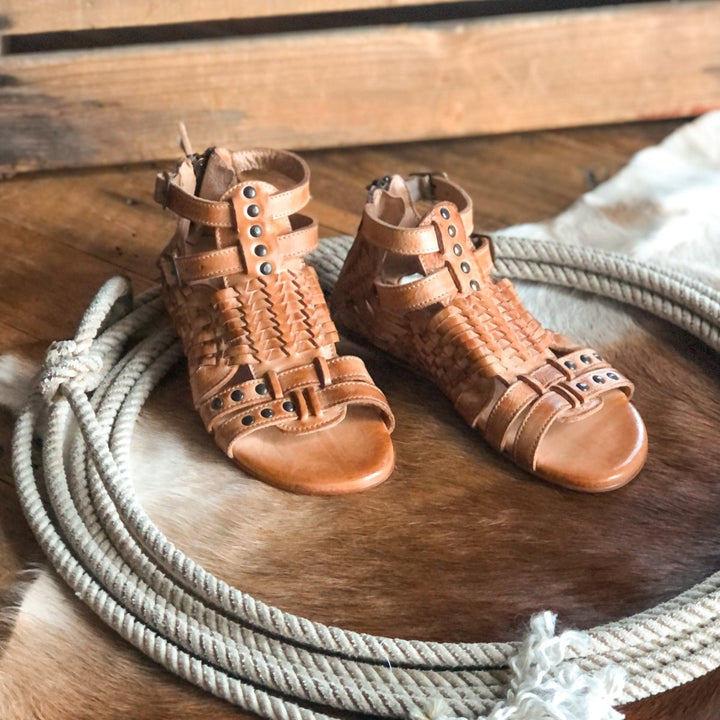 The Dacono tan woven leather flat sandals with metal studs by Savannah Sevens western life{&}style rest on a coiled rope, atop a cowhide rug and wooden floor. Sandals made by BedStu. 