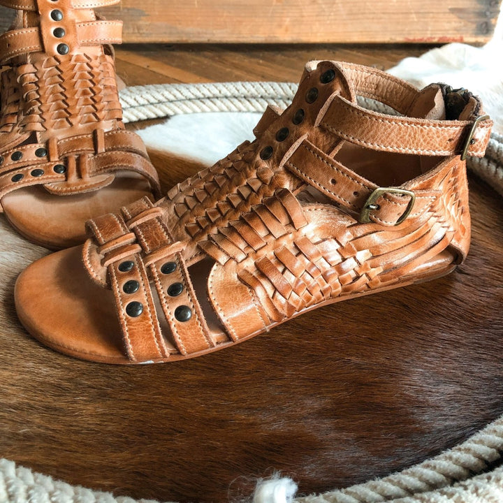 The Dacono sandals by Savannah Sevens western life{&}style feature comfy woven leather, ankle straps, and metal studs, showcased on a circular brown and white cowhide rug. Sandals made by BedStu. 