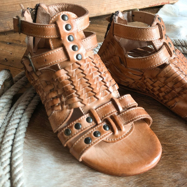 The Dacono by Savannah Sevens western life{&}style: tan woven leather sandals with open toes and metal stud accents, displayed on a cowhide rug with a rope nearby. Sandals made by BedStu. 