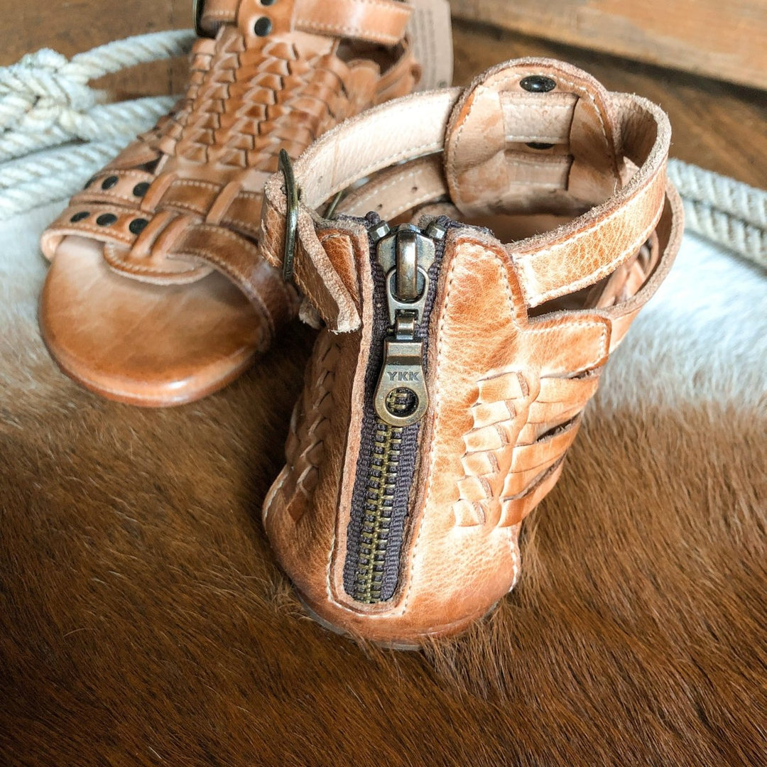 A close-up of The Dacono woven brown leather sandal by Savannah Sevens western life{&}style features a bold front zipper, resting on fur with another comfortable leather sandal partially visible in the background. Sandals made by BedStu. 