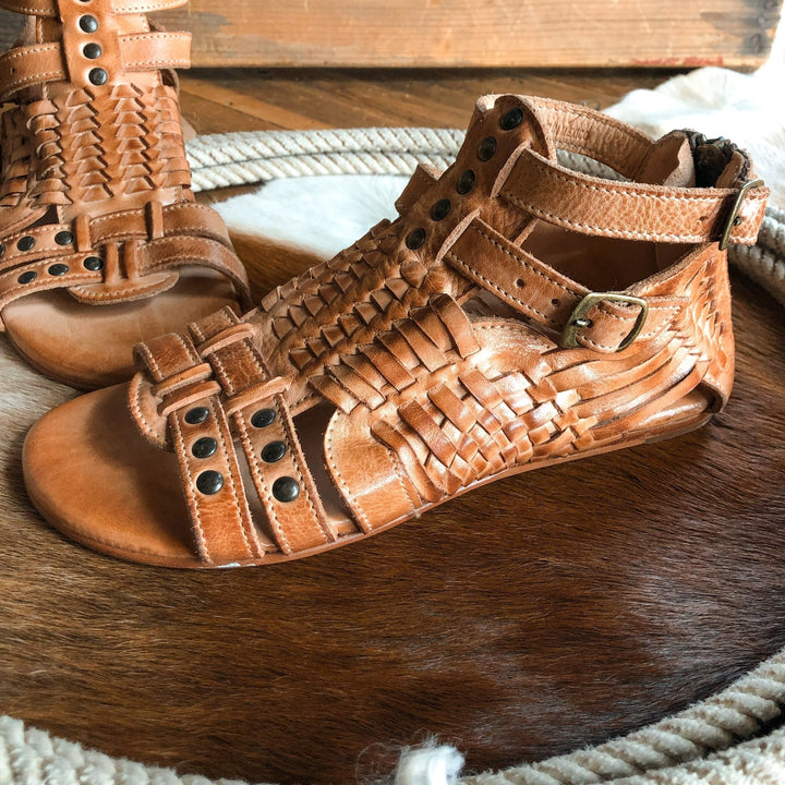 The Dacono sandals by Savannah Sevens western life{&}style feature comfy woven leather, ankle straps, and metal studs, showcased on a circular brown and white cowhide rug. Sandals made by BedStu. 
