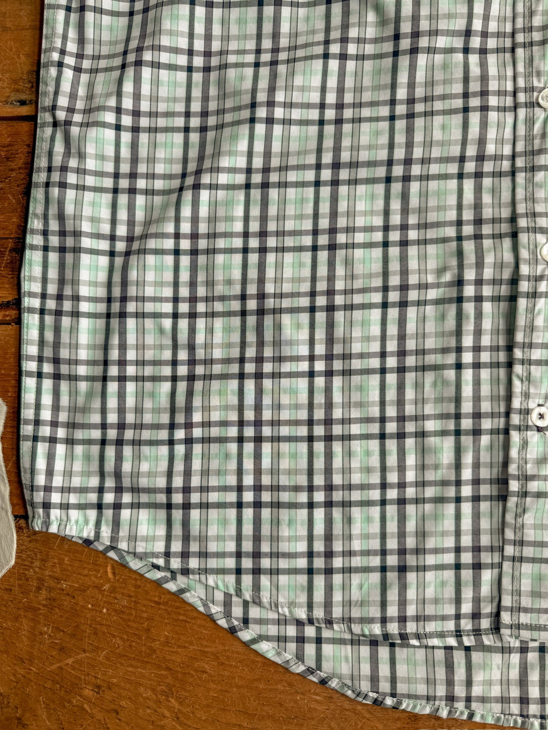A close-up of the Danville Cinch Men's Plaid Button Up by Savannah Sevens western life{&}style, featuring black, green, and light blue plaid on white; partially buttoned and flat on wood. Moisture wicking adds comfort.