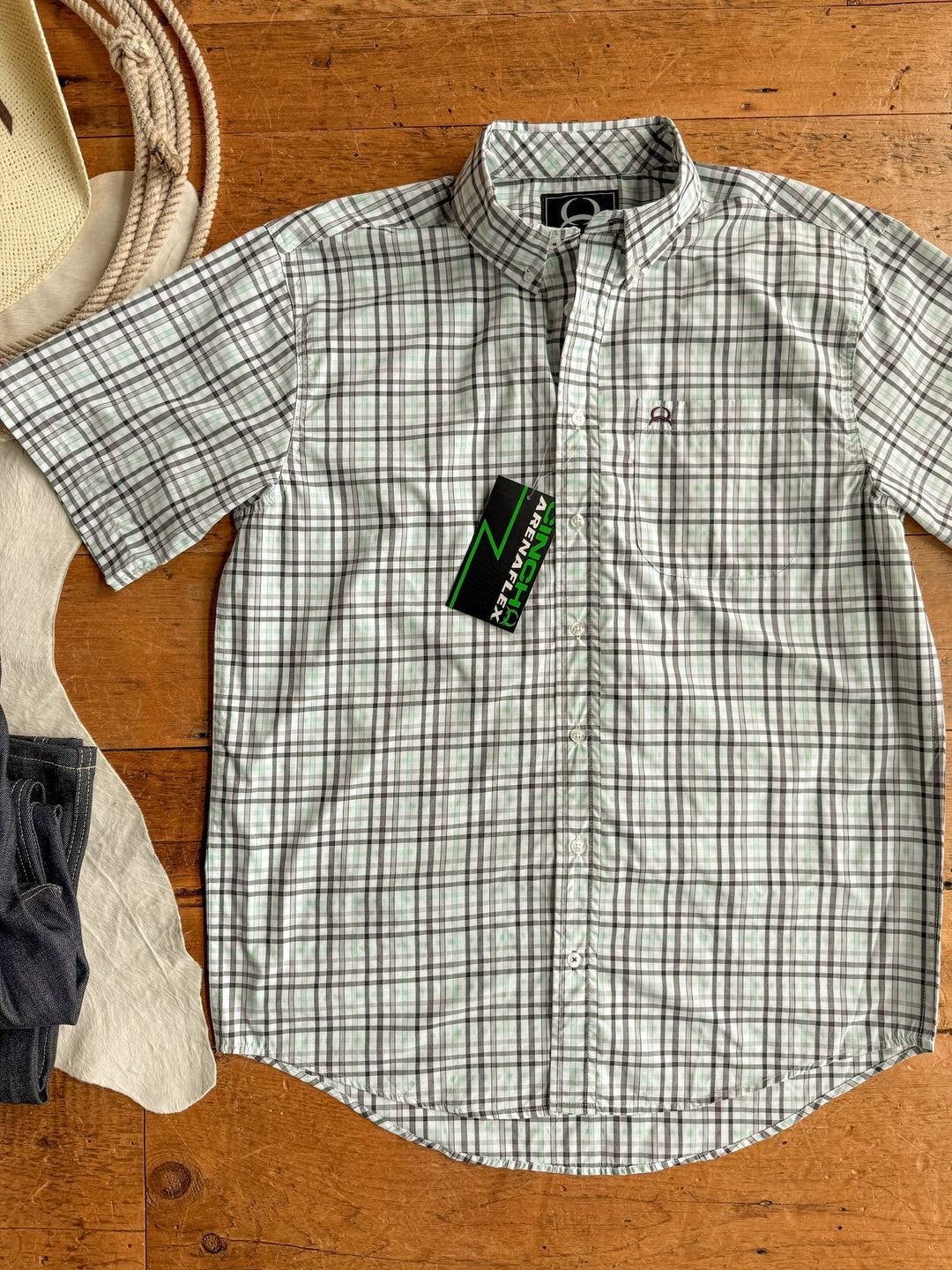 The Danville Cinch Men's Plaid Button Up by Savannah Sevens western life{&}style in green and white is laid flat on wood with a green and black tag. Cowboy hat, white fabric, and folded dark jeans appear on the left. Features moisture-wicking comfort.