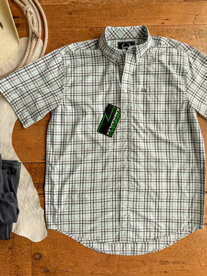 The Danville Cinch Men's Plaid Button Up by Savannah Sevens western life{&}style in green and white is laid flat on wood with a green and black tag. Cowboy hat, white fabric, and folded dark jeans appear on the left. Features moisture-wicking comfort.