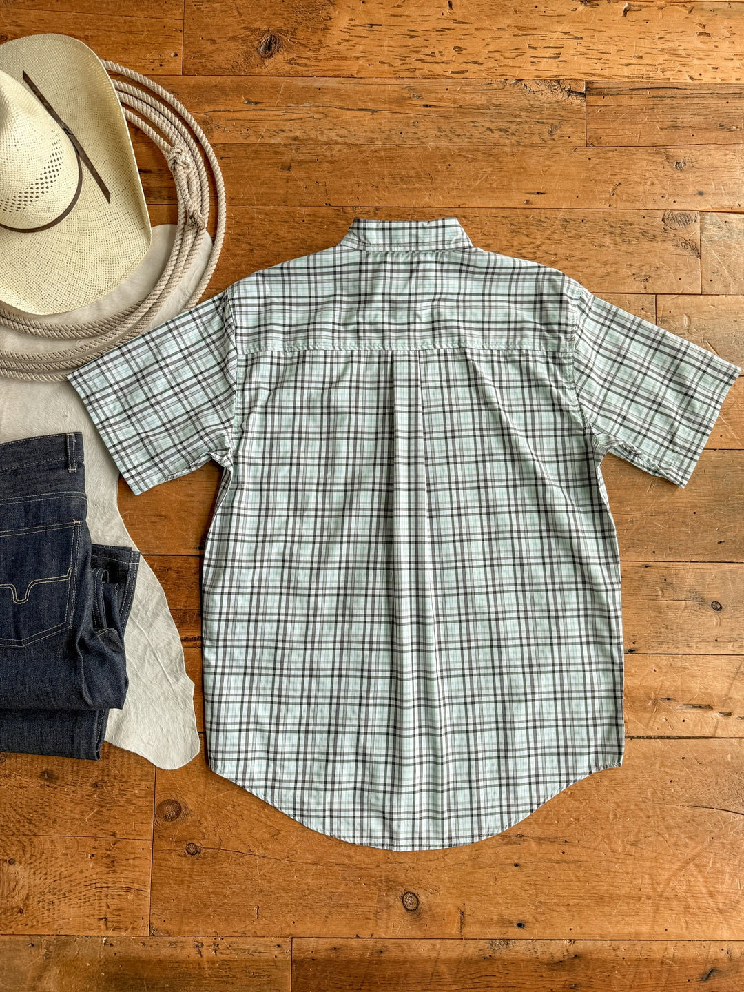 The Danville Cinch Men's Plaid Button Up by Savannah Sevens western life{&}style is laid flat, back up, on a wooden floor with a cowboy hat, dark jeans, and light fabric. This moisture-wicking shirt delivers comfortable style.