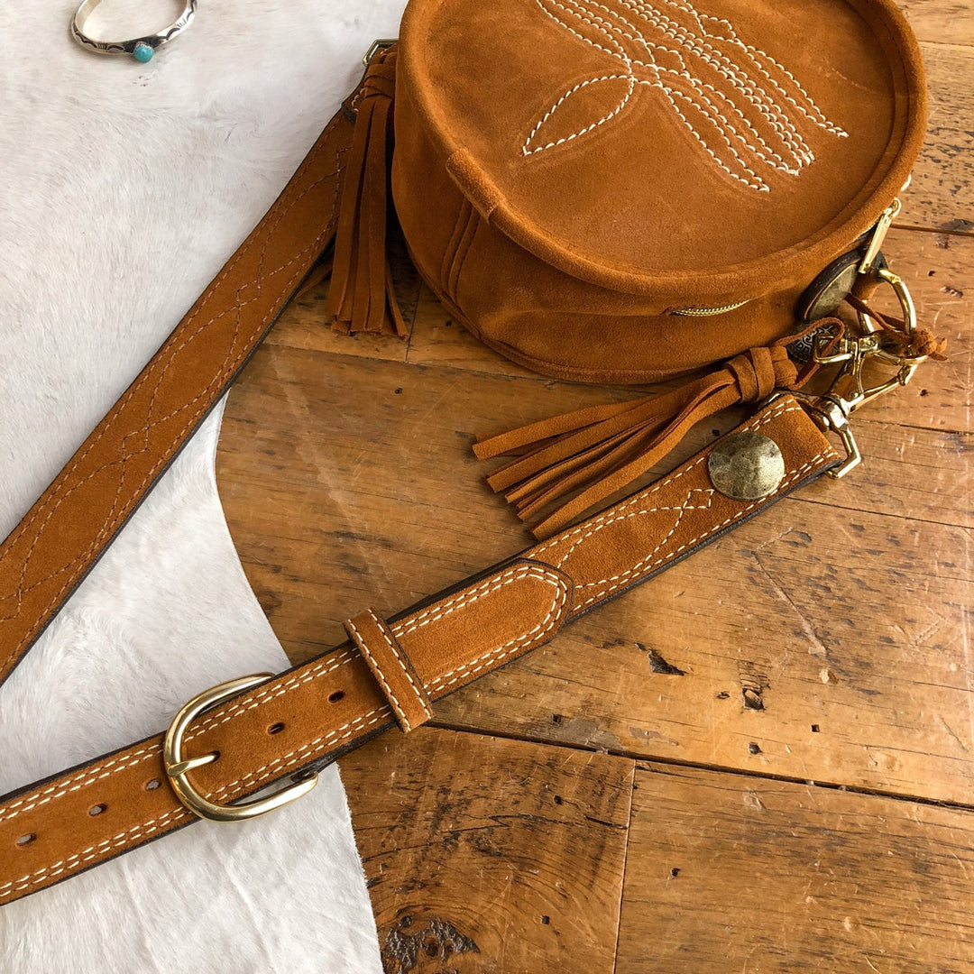 The Duvall by Savannah Sevens western life{&}style: a round brown suede crossbody bag with white stitching and tassel accents, shown on wood beside a matching brown leather belt with brass buckle. A silver ring is also partly visible. By Double J. 