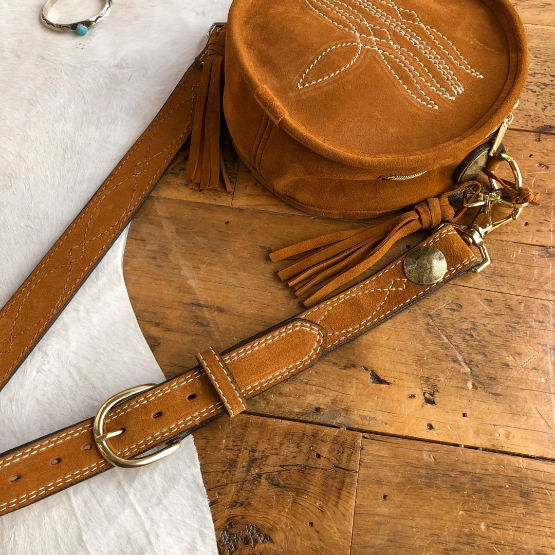 The Duvall by Savannah Sevens western life{&}style: a round brown suede crossbody bag with white stitching and tassel accents, shown on wood beside a matching brown leather belt with brass buckle. A silver ring is also partly visible. By Double J. 