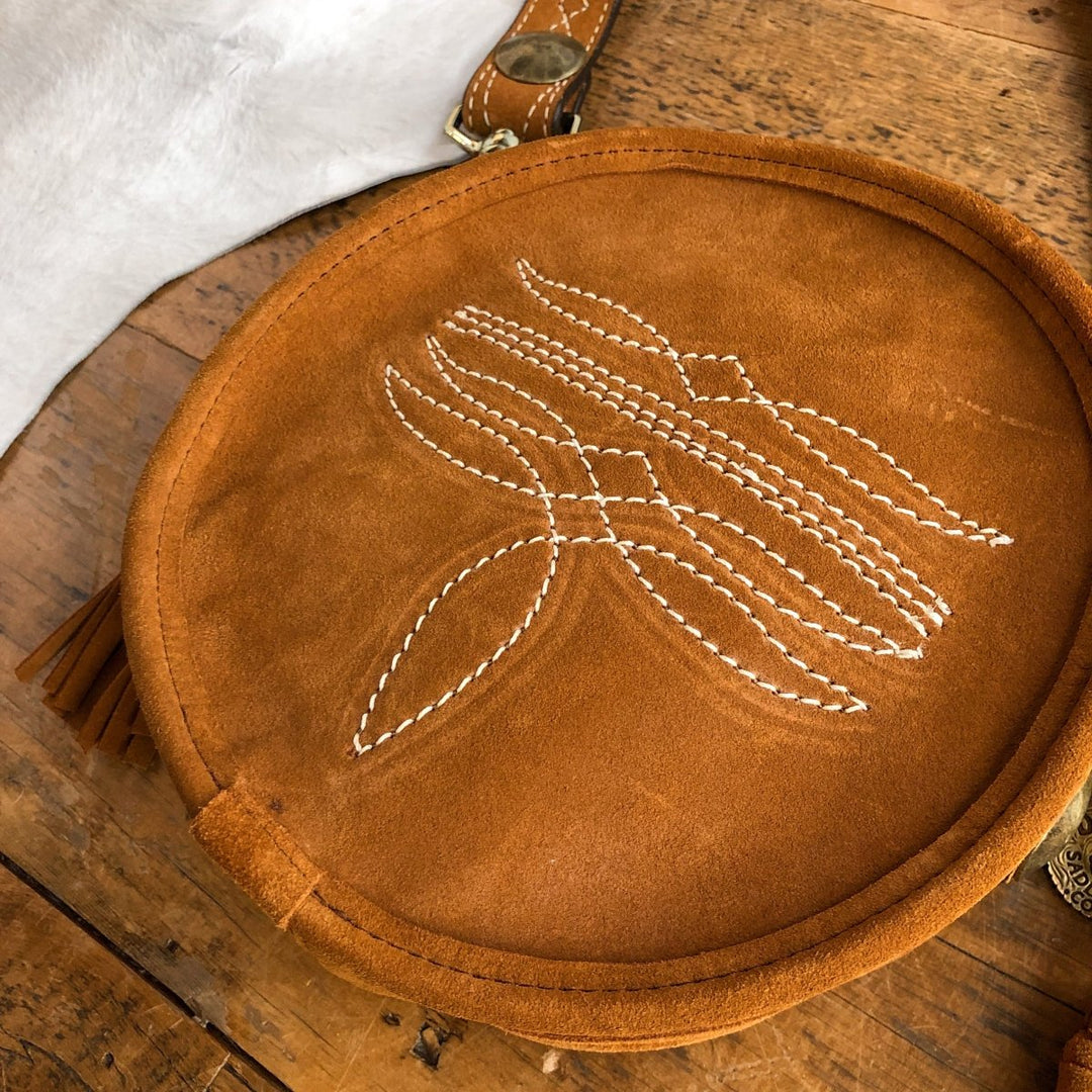 The Duvall by Savannah Sevens western life{&}style is a round brown suede handbag with white stitched patterns, accented by white fur and a brown leather strap with brass details, resting on a rustic wooden table. By Double J. 