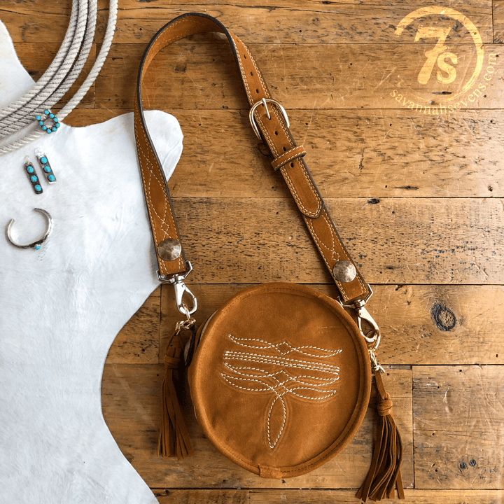 The Duvall, a round cognac leather handbag with white stitching and tassels from Savannah Sevens western life{&}style, is displayed on a wooden surface next to turquoise jewelry. The brand logo appears in the top right corner. By Double J. 
