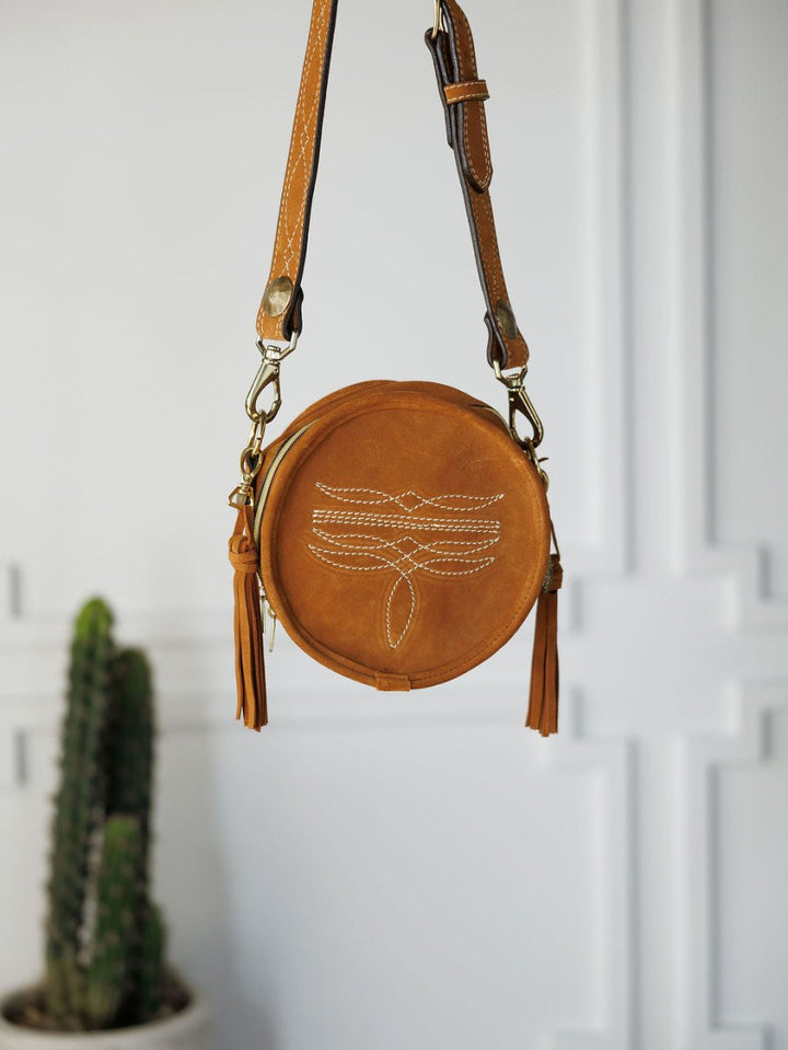 The Duvall by Savannah Sevens western life{&}style is a round cognac leather handbag with white stitching and tassels, hanging from its strap. A blurred green cactus in a pot sits in the background against a white wall.