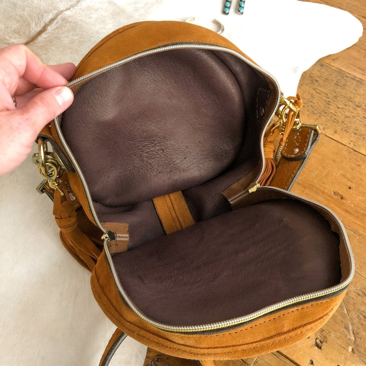 A hand opens The Duvall by Savannah Sevens western life{&}style, revealing its brown leather interior, main compartment, and zipper on a wooden surface. By Double J. 