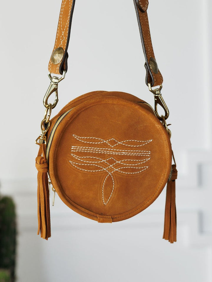 The Duvall by Savannah Sevens western life{&}style is a round tan leather handbag with white stitching, gold hardware, and tassel accents, shown against a white background.