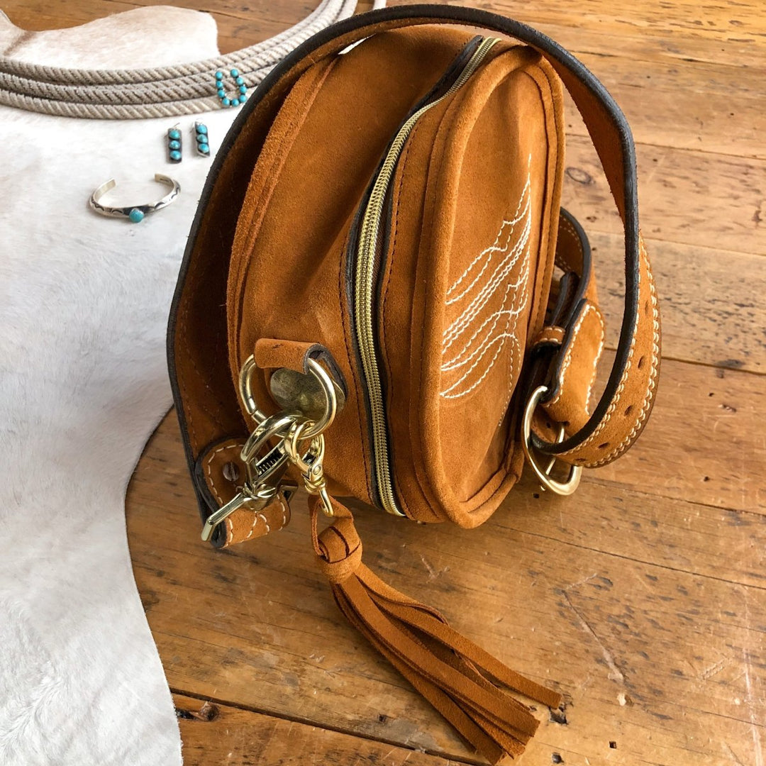 The Duvall by Savannah Sevens western life{&}style—a brown suede crossbody with white stitching, brass hardware, zipper, and leather tassel—rests on wood. Turquoise jewelry and a coiled rope lay nearby on a white hide. By Double J. 