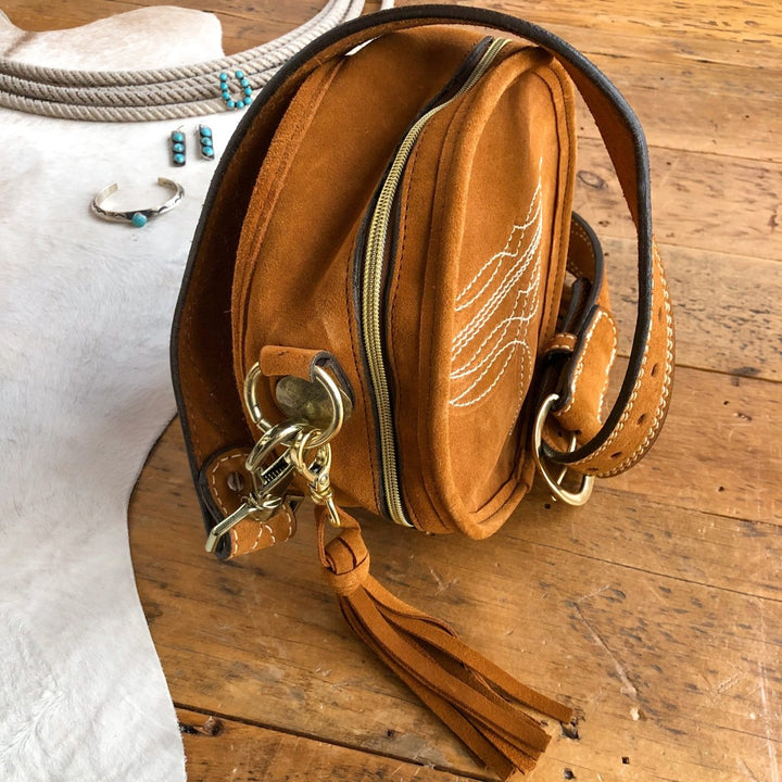 The Duvall by Savannah Sevens western life{&}style—a brown suede crossbody with white stitching, brass hardware, zipper, and leather tassel—rests on wood. Turquoise jewelry and a coiled rope lay nearby on a white hide. By Double J. 