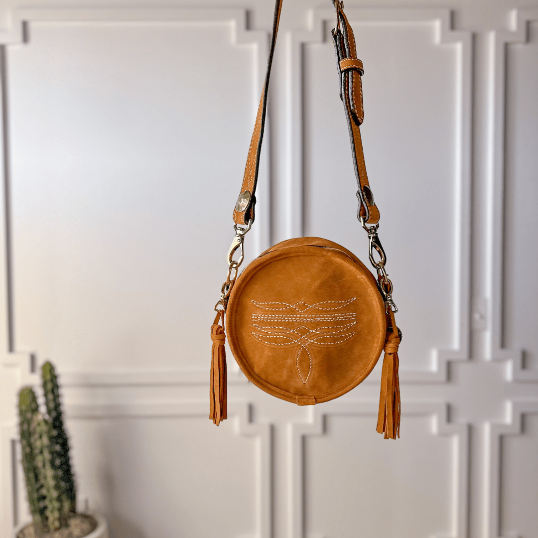 The Duvall, a round cognac leather handbag with tassels and an embroidered dragonfly by Savannah Sevens western life{&}style, hangs against a white paneled wall with a small potted cactus in the background. By Double J. 