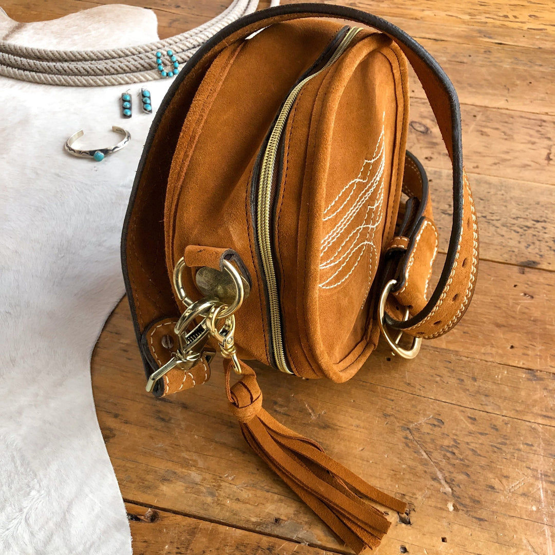 The Duvall by Savannah Sevens western life{&}style—a brown suede crossbody with white stitching, brass hardware, zipper, and leather tassel—rests on wood. Turquoise jewelry and a coiled rope lay nearby on a white hide. By Double J. 