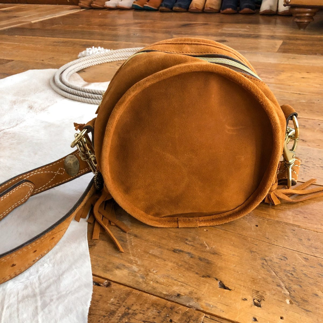 The Duvall, a round cognac leather handbag by Savannah Sevens western life{&}style with a gold zipper and fringe, is placed on a wooden floor beside a white rug and coiled rope. By Double J. 