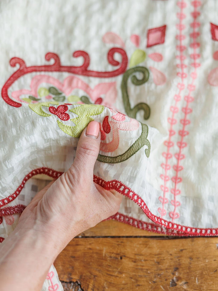 A hand holds the edge of The Elrosa Set by Savannah Sevens western life{&}style—a white floral embroidered top and shorts set with red, pink, and green designs—resting on wood.