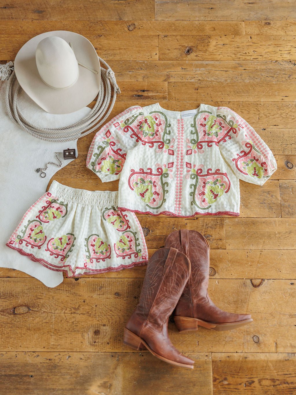 The Elrosa {Set} by Savannah Sevens western life{&}style—a cream floral embroidered top, high waisted shorts, —lays on a wooden floor with brown cowboy boots, a wide-brimmed hat, and a cowhide rug.