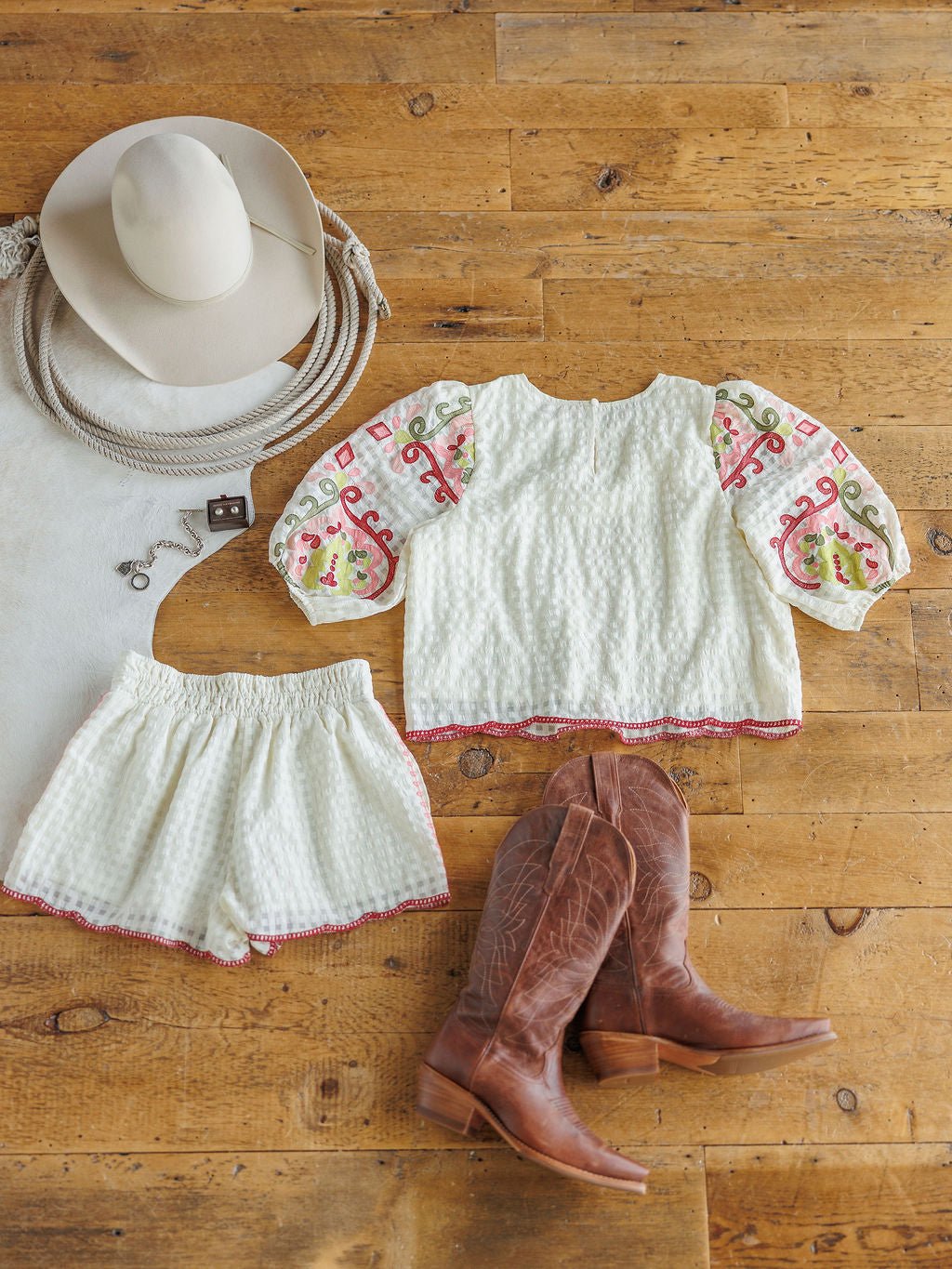 The Elrosa {Set} by Savannah Sevens western life{&}style is styled on a wooden floor with its floral embroidered top and shorts, colorful sleeves, high waisted shorts, brown cowboy boots, a white cowboy hat, rope, and a bold buckle belt.