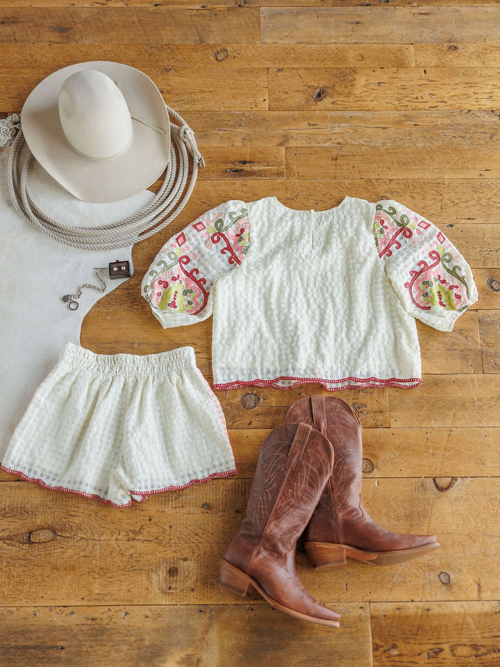The Elrosa {Set} by Savannah Sevens western life{&}style is styled on a wooden floor with its floral embroidered top and shorts, colorful sleeves, high waisted shorts, brown cowboy boots, a white cowboy hat, rope, and a bold buckle belt.