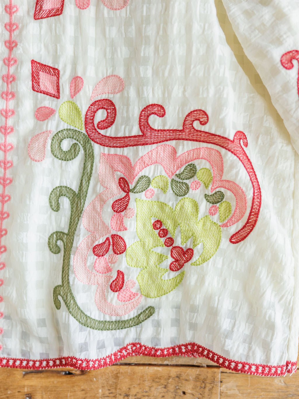 A close-up of The Elrosa {Set} from Savannah Sevens western life{&}style shows white fabric with vibrant floral and swirl embroidery in green, red, and pink, finished with red trim—suggesting a chic embroidered top & shorts set on wood.