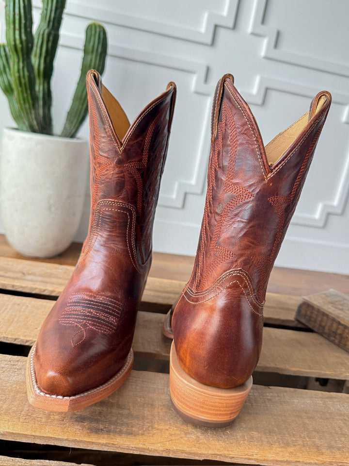 The Emerado chestnut leather snip toe cowboy boots by Savannah Sevens western life{&}style feature detailed stitching and sit on a wooden surface, with a green cactus in a white pot and a textured white wall in the background. Made by Black Star. 