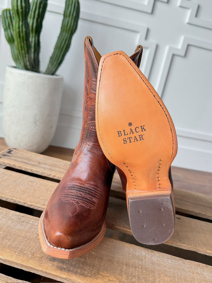 A pair of The Emerado chestnut leather cowboy boots by Savannah Sevens western life{&}style—one upright, one tilted to reveal the BLACK STAR-stamped sole—rest on wood with a potted cactus in the background. Made by Black Star. 