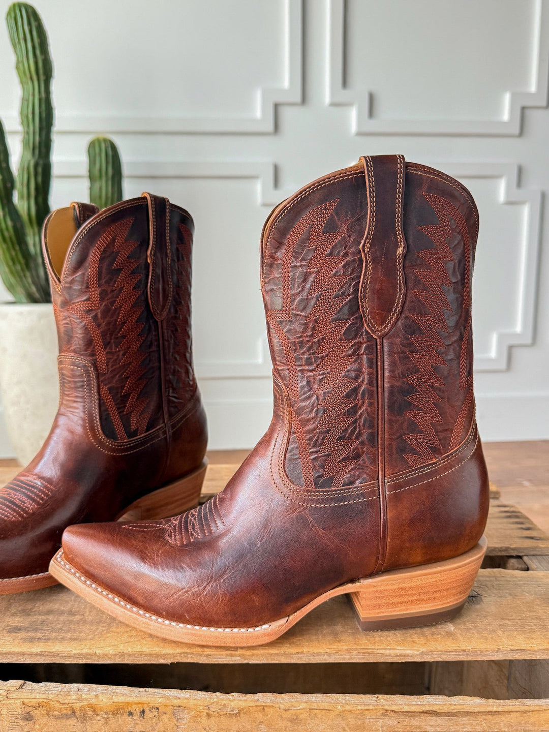 The Emerado chestnut leather cowboy boots by Savannah Sevens western life{&}style, featuring decorative stitching and a snip toe, rest on wood with a cactus in a white pot and a white paneled wall in the background. Made by Black Star. 