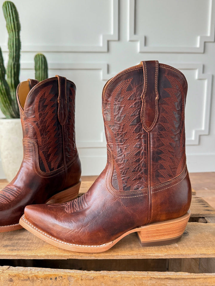 The Emerado chestnut leather cowboy boots by Savannah Sevens western life{&}style, featuring decorative stitching and a snip toe, rest on wood with a cactus in a white pot and a white paneled wall in the background. Made by Black Star. 