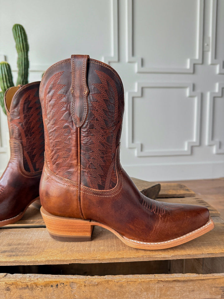 The Emerado cowboy boots by Savannah Sevens western life{&}style feature chestnut leather, decorative stitching, a snip toe, and a light stacked heel. They sit on wood with a green cactus and white paneled walls in the background. Made by Black Star. 
