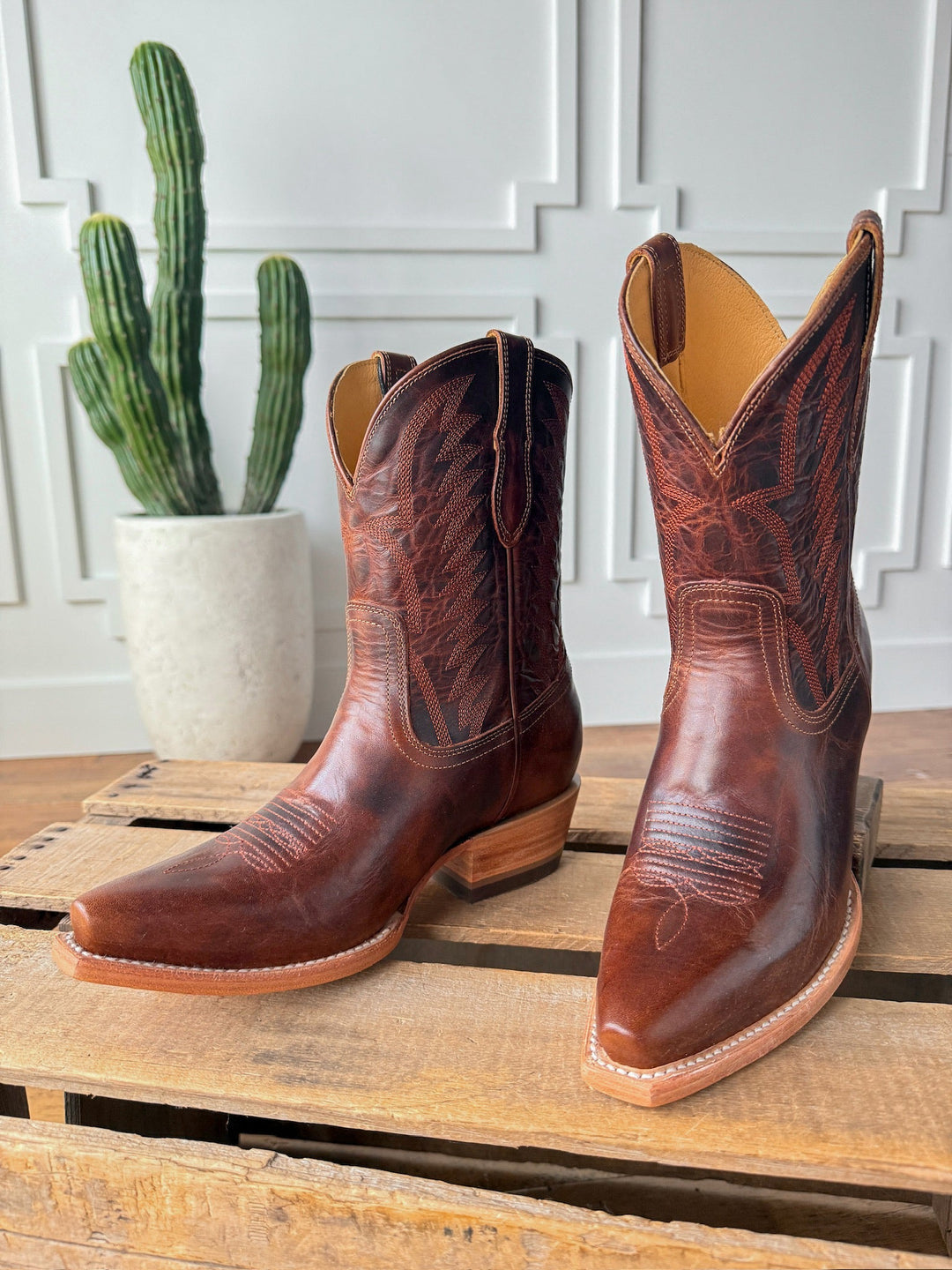 The Emerado chestnut leather cowboy boots by Savannah Sevens western life{&}style, featuring decorative stitching and a snip toe, are displayed on a wooden pallet with a potted cactus and white paneled wall in the background. Made by Black Star. 