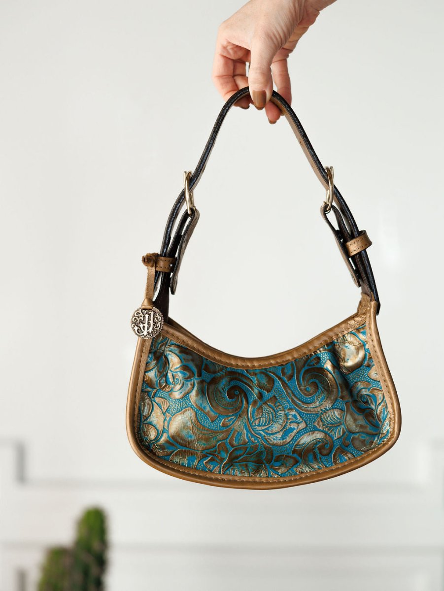 A hand displays The Ganado handbag by Savannah Sevens western life{&}style, featuring a blue and gold swirling design, tan trim, black handles, and a round metallic charm reminiscent of turquoise tooled leather.
