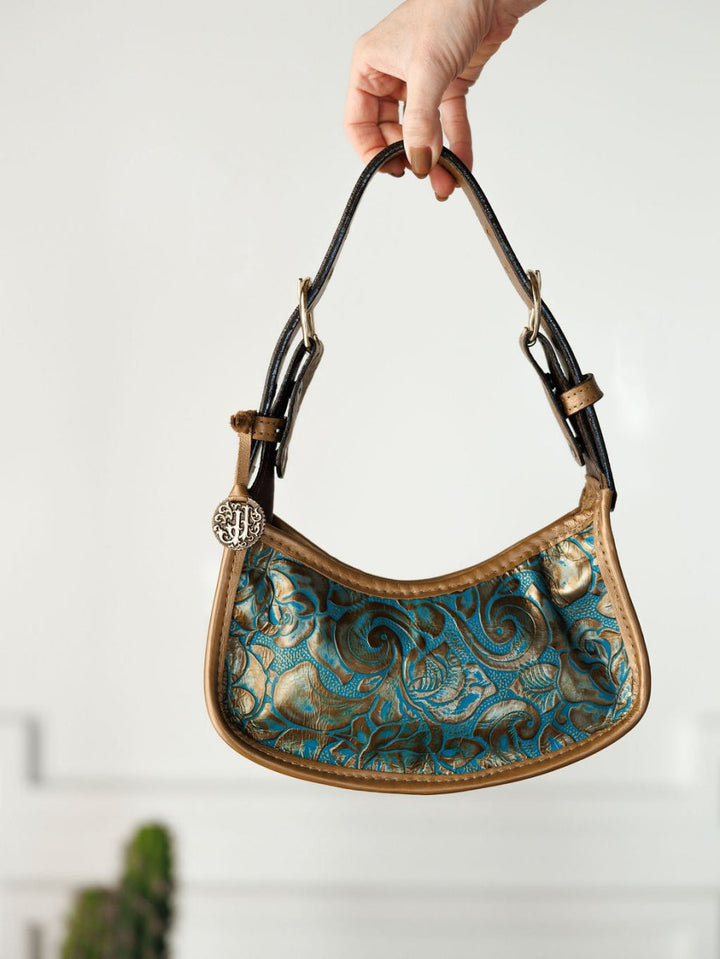 A hand displays The Ganado handbag by Savannah Sevens western life{&}style, featuring a blue and gold swirling design, tan trim, black handles, and a round metallic charm reminiscent of turquoise tooled leather.