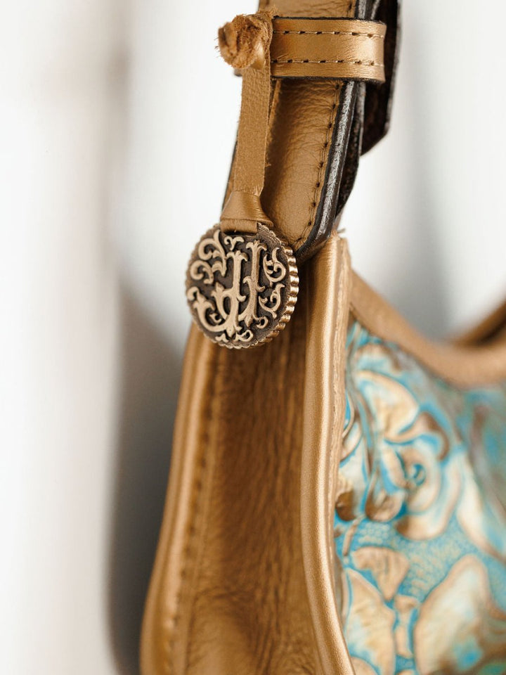 Close-up of The Ganado handbag by Savannah Sevens western life{&}style, showcasing hand-tooled leather, a decorative metallic button with ornate floral design, and blue and gold floral embroidery.