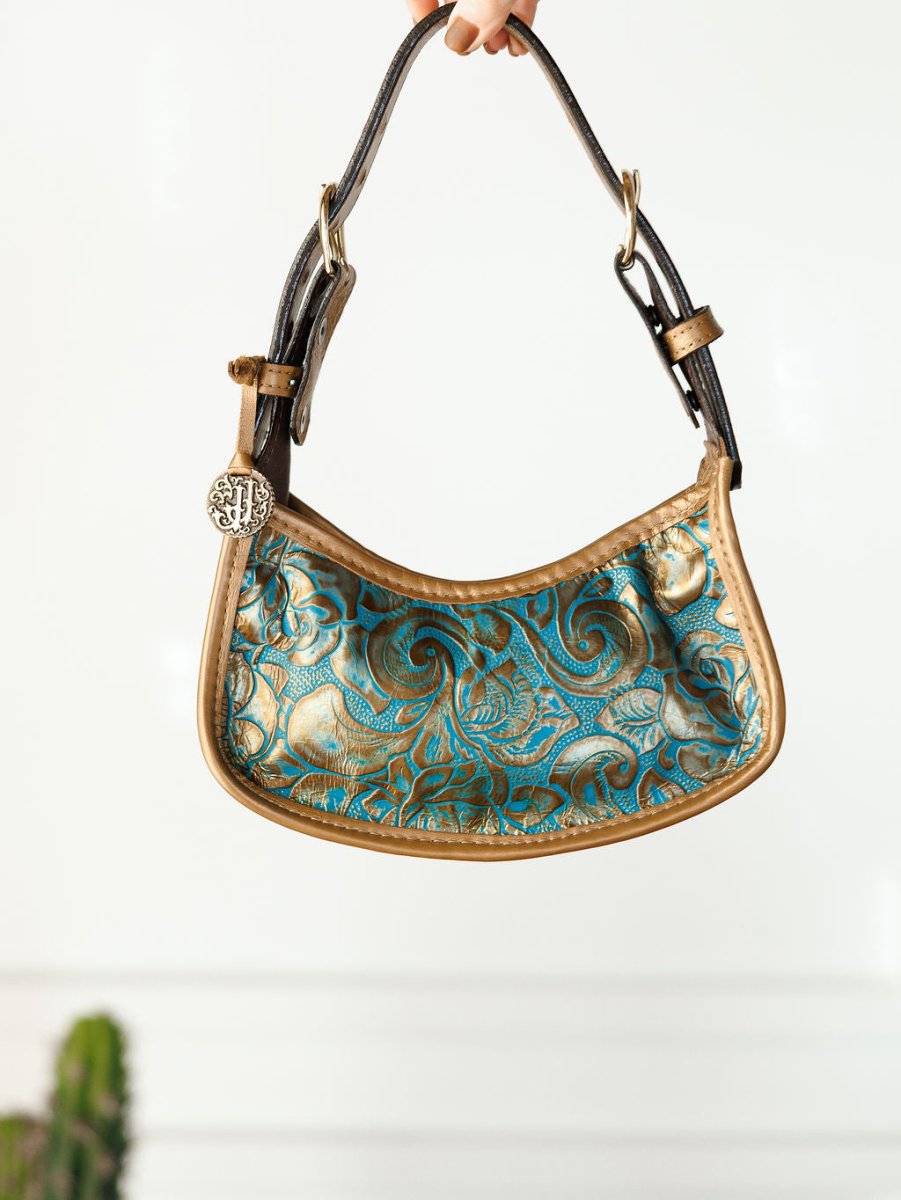 A hand holds The Ganado handbag by Savannah Sevens western life{&}style, featuring brown leather trim, blue and gold floral pattern, turquoise tooled leather accents, and a round metallic charm. A blurred plant appears in the background.