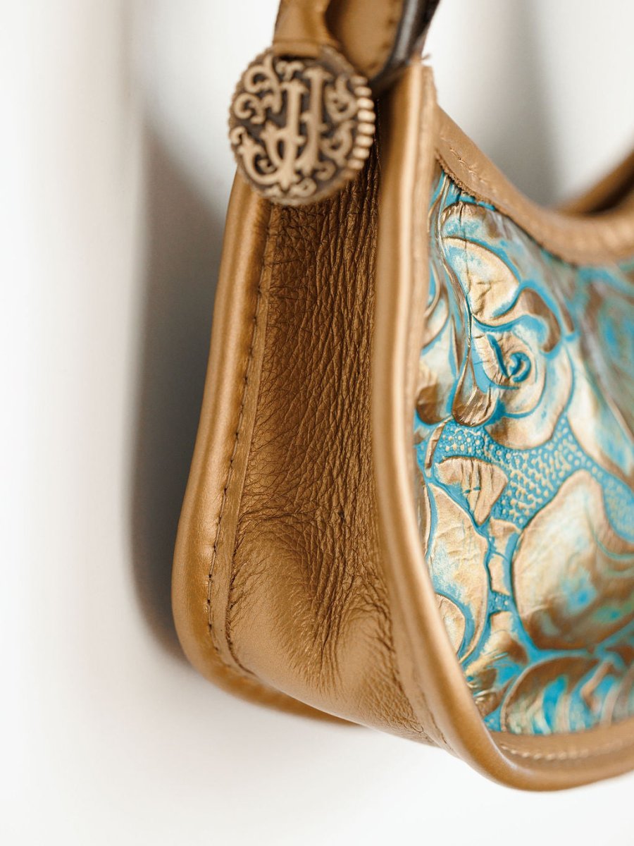 Close-up of The Ganado by Savannah Sevens western life{&}style, a hand tooled leather handbag with turquoise and gold floral embossing and a decorative metal button at the strap attachment.