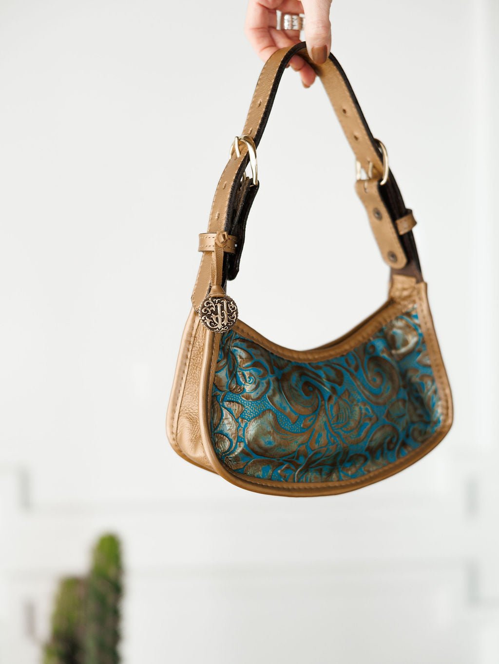 A hand holds The Ganado handbag by Savannah Sevens western life{&}style, featuring tan and turquoise tooled leather with an embossed floral pattern, black accents, and a round metallic charm. A blurred green plant appears against the white background.
